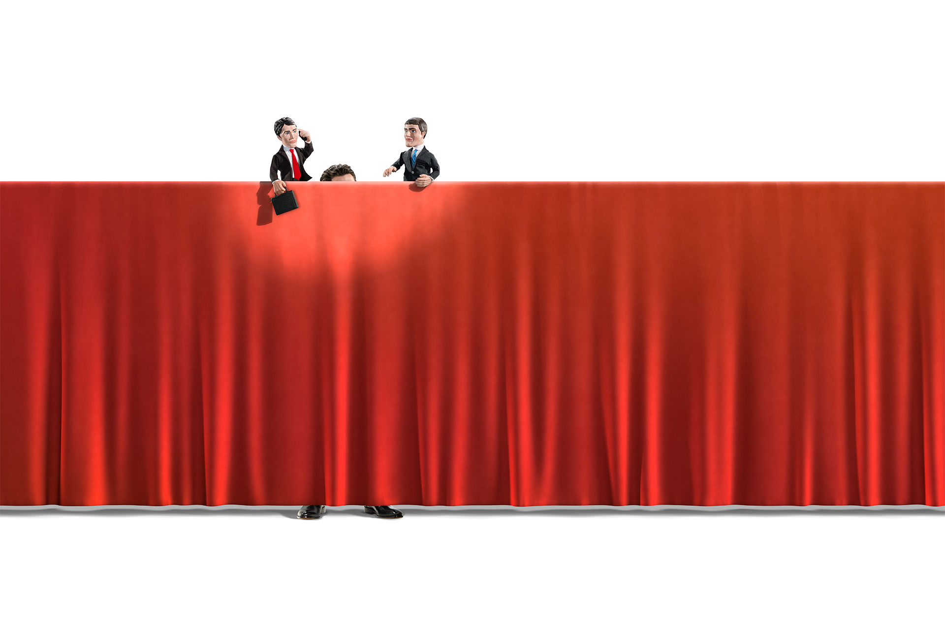 A wide red curtain serves as a stage for a Punch and Judy Theatre. A man standing behind the curtain operates two puppets looking like businessmen.